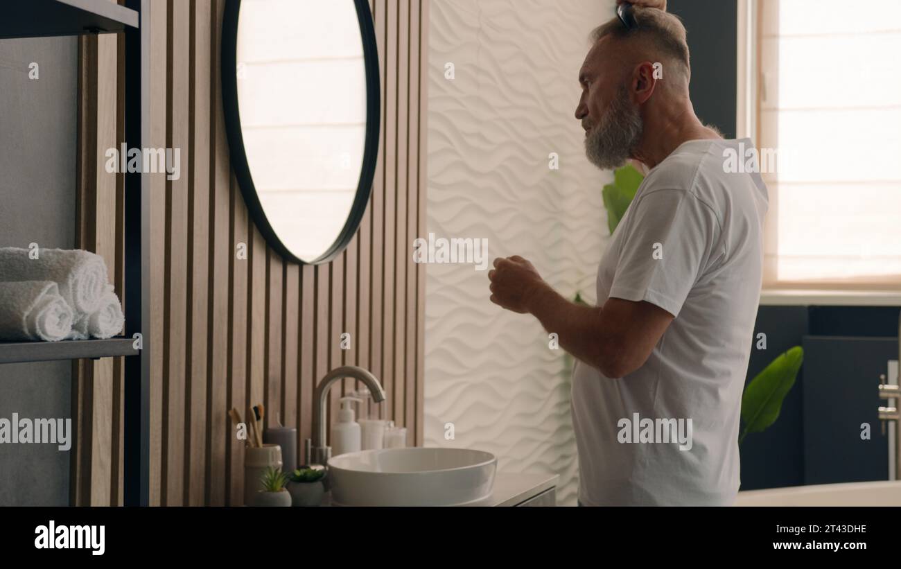 Beau vieil homme caucasien confiant dans la salle de bain regardant le reflet miroir peignant les cheveux de peigne de barbe avec brosse senior mature mâle brossant gris Banque D'Images