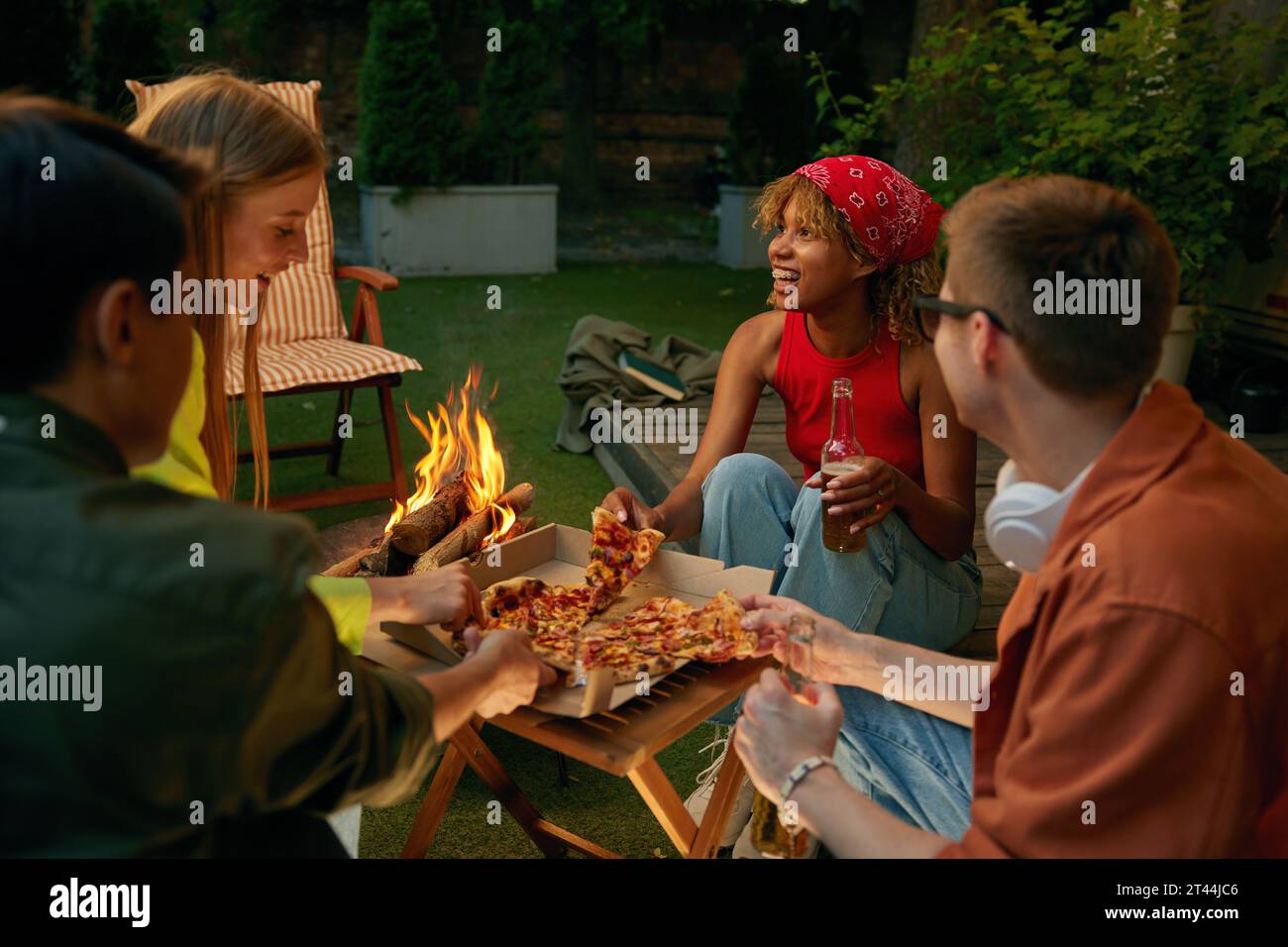 Heureux amis millénaires appréciant pizza et bière boire à proximité feu de camp Banque D'Images