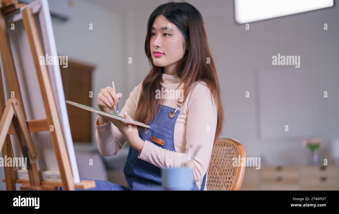 Jeune artiste féminine asiatique utilisant le pinceau pour mélanger la couleur sur la palette pour créer de la peinture sur toile. Banque D'Images