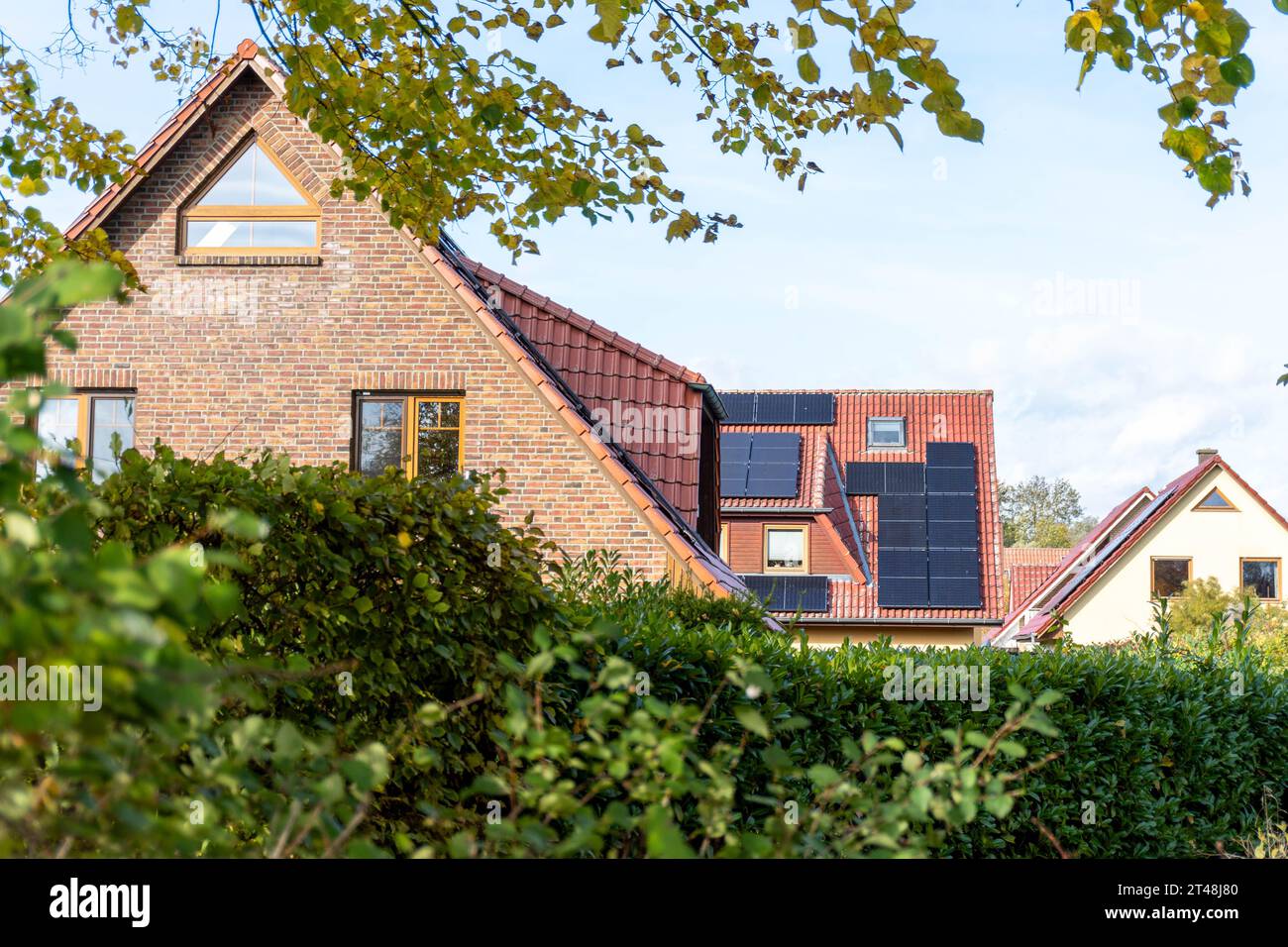maison en brique avec tuiles rouges et panneaux solaires se trouve derrière des haies luxuriantes dans un quartier paisible et verdoyant de banlieue par une journée ensoleillée Banque D'Images