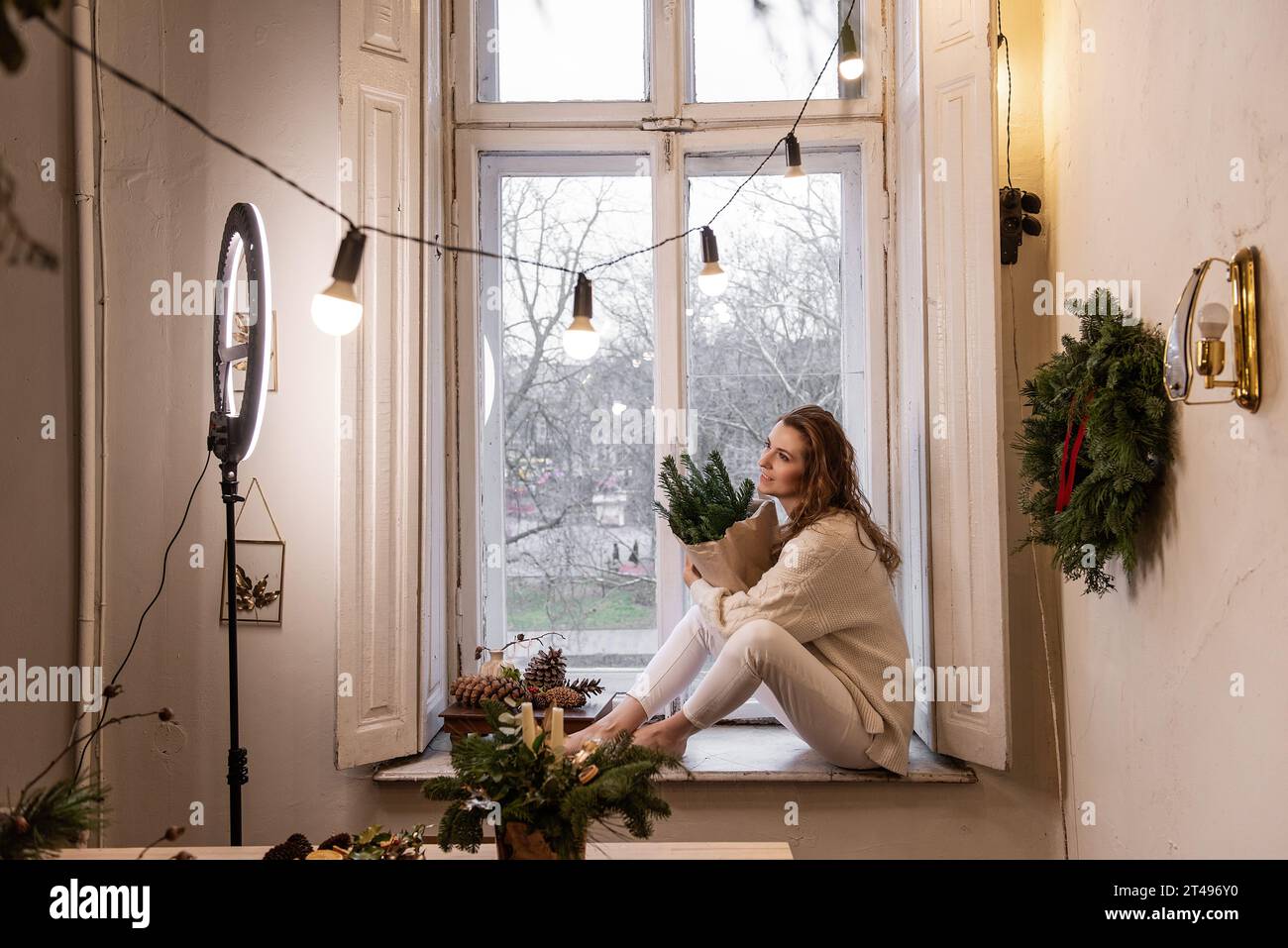 Jeune femme blogueuse est assise sur le rebord de la fenêtre près de la fenêtre, tient dans les mains bouquet de branches d'épinette verte dans du papier artisanal. Lampe LED à lumière annulaire, fleuriste fille Banque D'Images