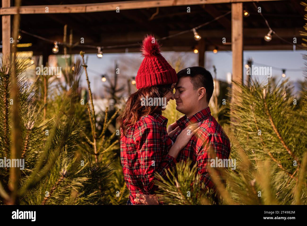 Tendres câlins de jeune couple amoureux parmi le marché vert des arbres de Noël en ville. Belle femme embrasse passionnément, regarde l'homme. Les jeunes mariés regardent Banque D'Images