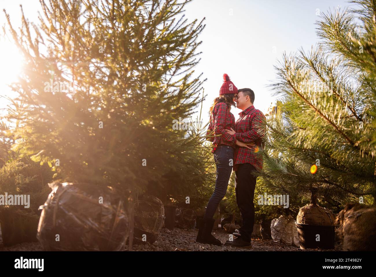 Joyeux couple amoureux dans des chemises rouges à carreaux, chapeaux tricotés se trompent, riant parmi le marché vert de l'arbre de Noël. Le jeune homme tient les mains de Banque D'Images