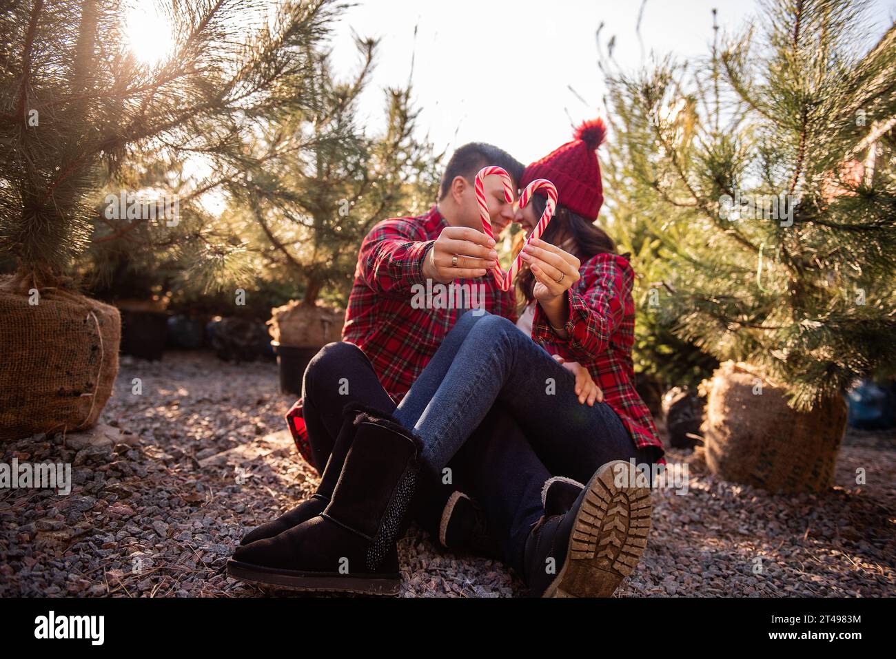 Couple amoureux en chemises à carreaux, chapeaux tricotés tiennent des cannes de bonbons en forme de cœur dans les mains près du marché vert des arbres de Noël. L'homme et la femme s'embrassent, rient, h. Banque D'Images
