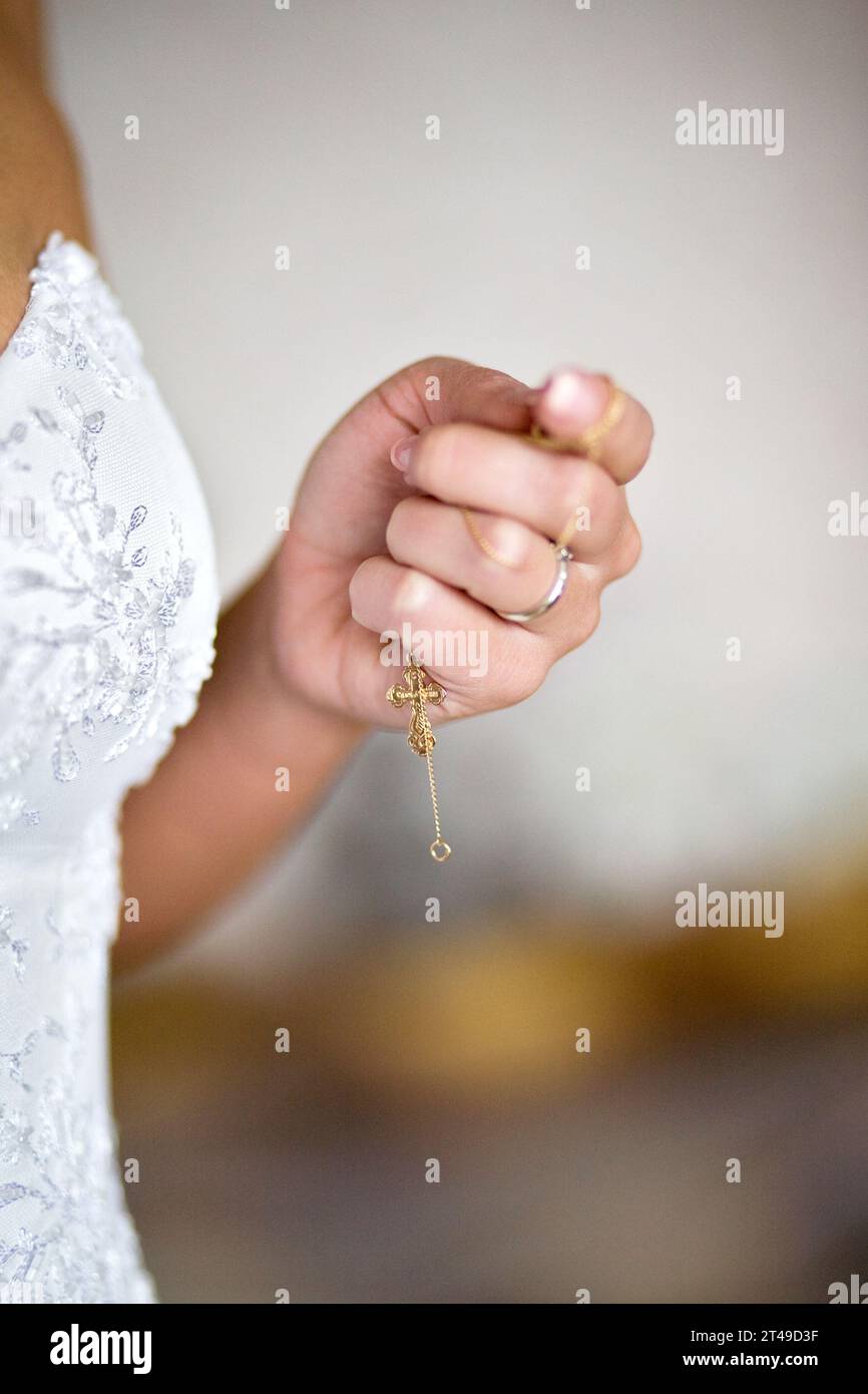Gros plan de la main d'une femme tenant un pendentif en croix dorée, avec une robe de mariée florale blanche en toile de fond. Banque D'Images