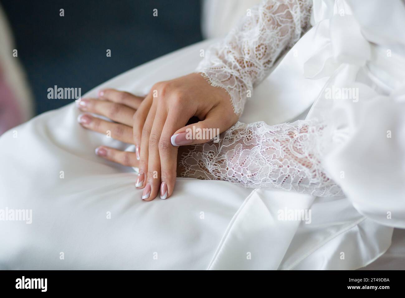 Gros plan des mains de la mariée avec des ongles roses sur une robe blanche, soulignée par des manches en dentelle dans une lumière douce. Banque D'Images
