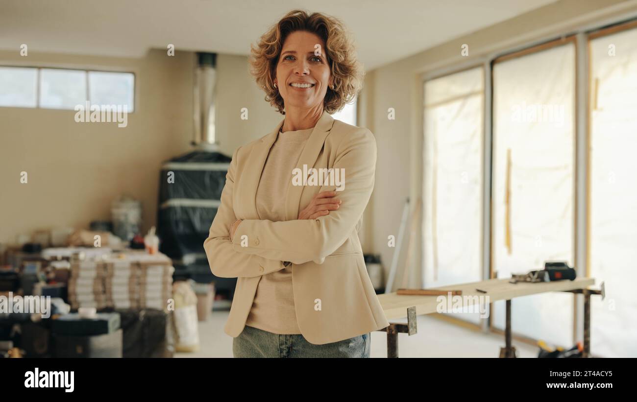Femme heureuse et professionnelle se tient dans une maison en cours de rénovation les bras croisés. Elle est gestionnaire de projet et propriétaire, supervisant le remo Banque D'Images