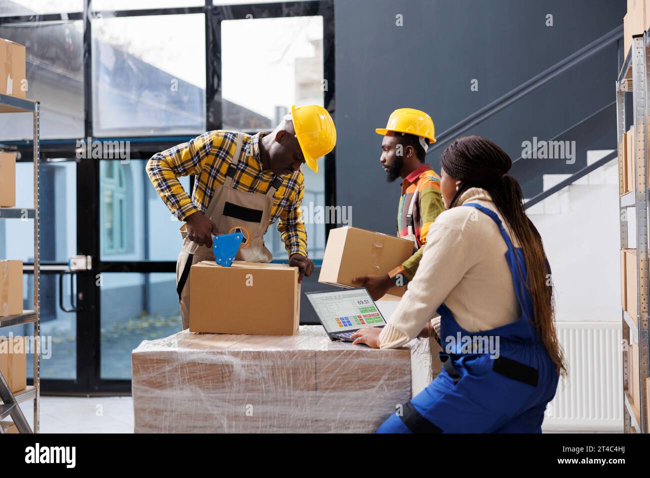Équipe de gestionnaires d'entrepôt afro-américains préparant la commande du client pour la livraison. Tous les employés de magasin noir emballant la boîte en carton avec du ruban adhésif et vérifiant la liste des marchandises sur l'ordinateur portable Banque D'Images