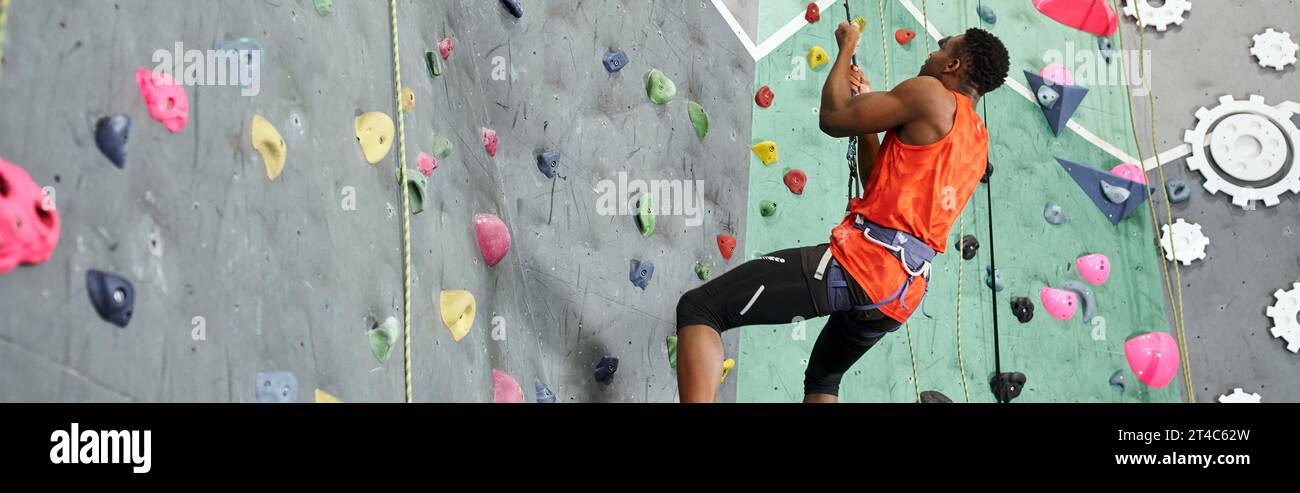 homme afro-américain musclé en chemise orange accroché à sa corde d'escalade et regardant vers le haut, bannière Banque D'Images