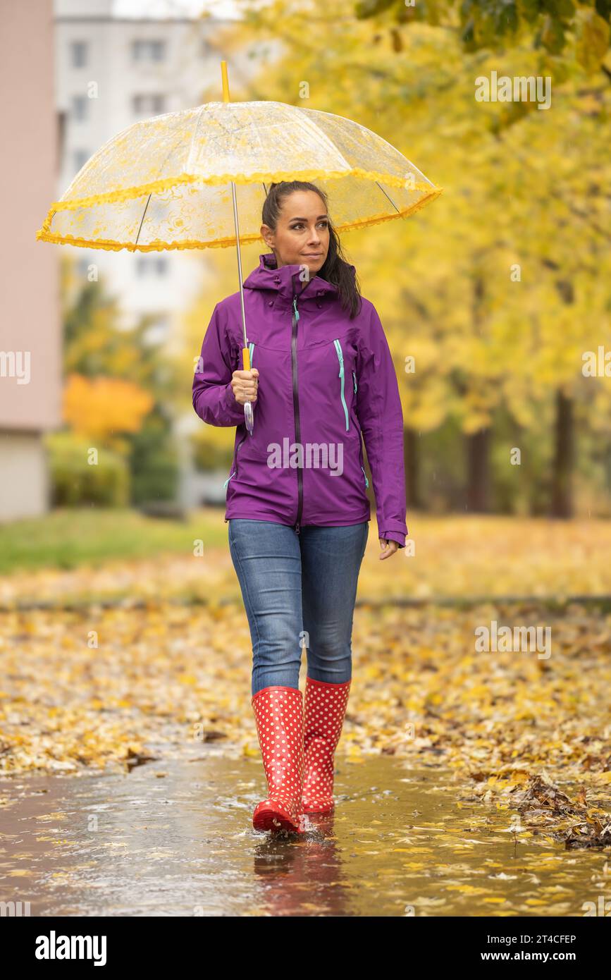 Une belle brune dans des vêtements chauds et avec un parapluie se promène dans un parc de la ville entouré de feuilles d'automne tombées. Banque D'Images