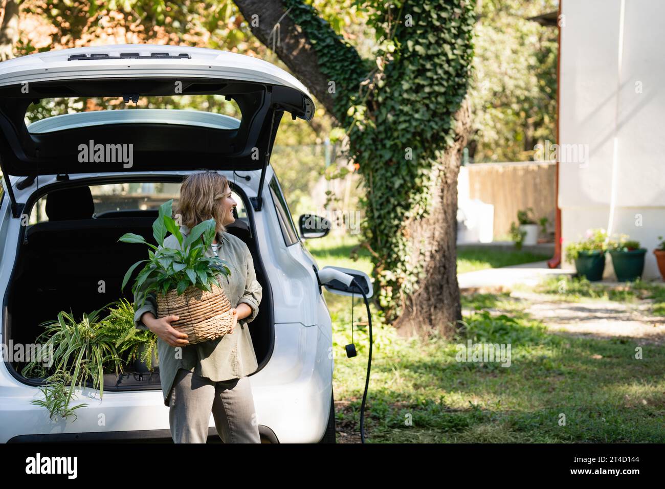 Femme avec plante en pot à côté d'une voiture électrique de charge dans la cour d'une maison de campagne. Banque D'Images