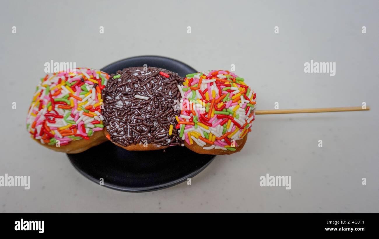 beignets avec des saupoudrages colorés et des saupoudrages de chocolat sur des brochettes comme satay sur une assiette noire Banque D'Images