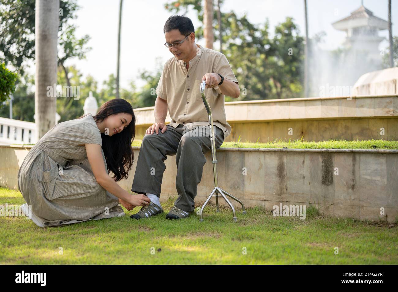 Une jeune fille asiatique adorable et attentionnée aide son père aîné à mettre des chaussures, nouer des lacets tout en se relaxant dans un parc verdoyant à l'extérieur. Banque D'Images