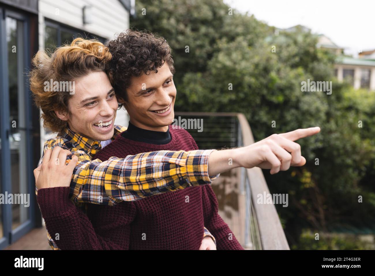Heureux et diversifié couple masculin gay embrassant et pointant du doigt au balcon sur la vue de la forêt Banque D'Images