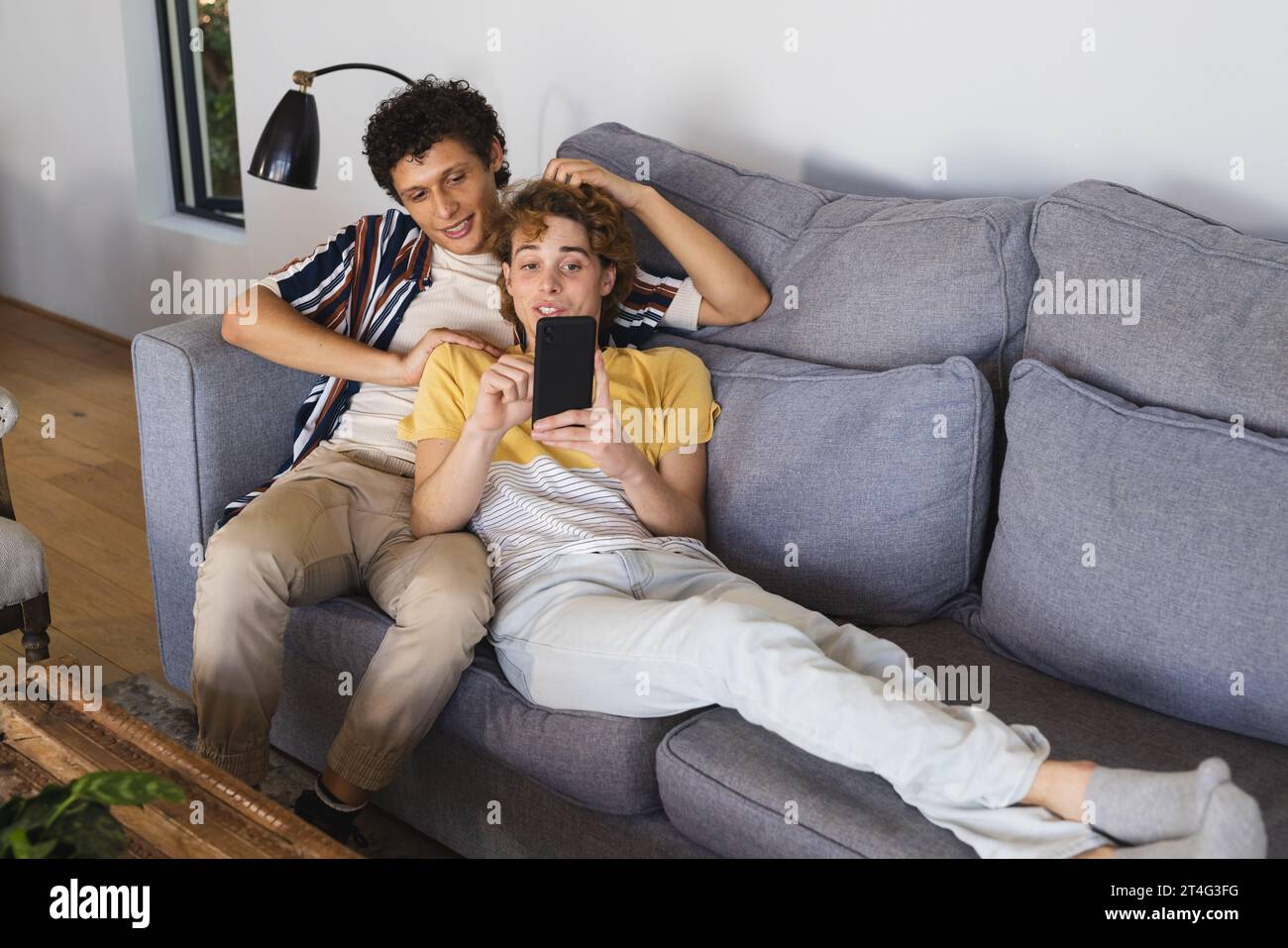 Heureux couple masculin gay diversifié utilisant smartphone et embrassant à la maison Banque D'Images