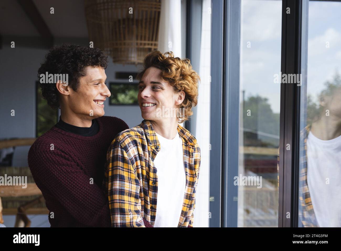 Heureux et diversifié couple masculin gay embrassant et se regardant à la fenêtre à la maison Banque D'Images