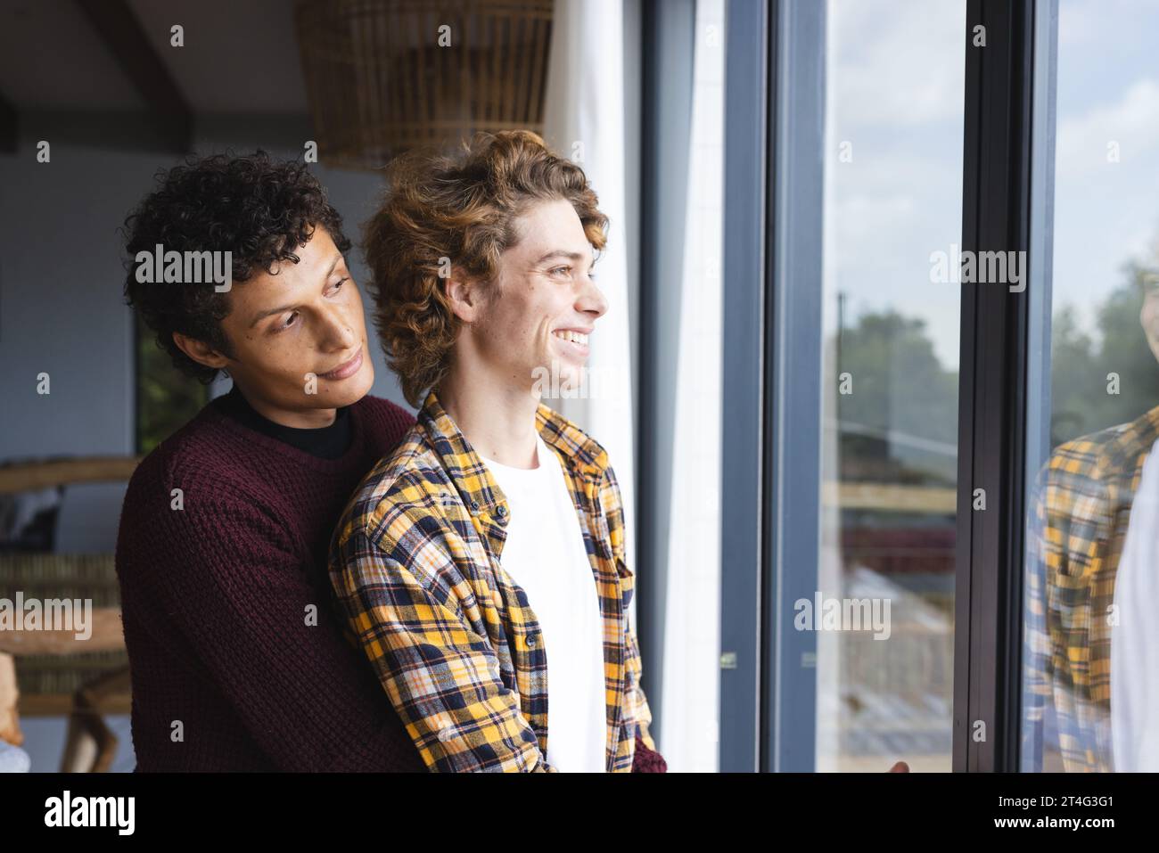 Heureux couple masculin gay diversifié embrassant et regardant par la fenêtre à la maison Banque D'Images