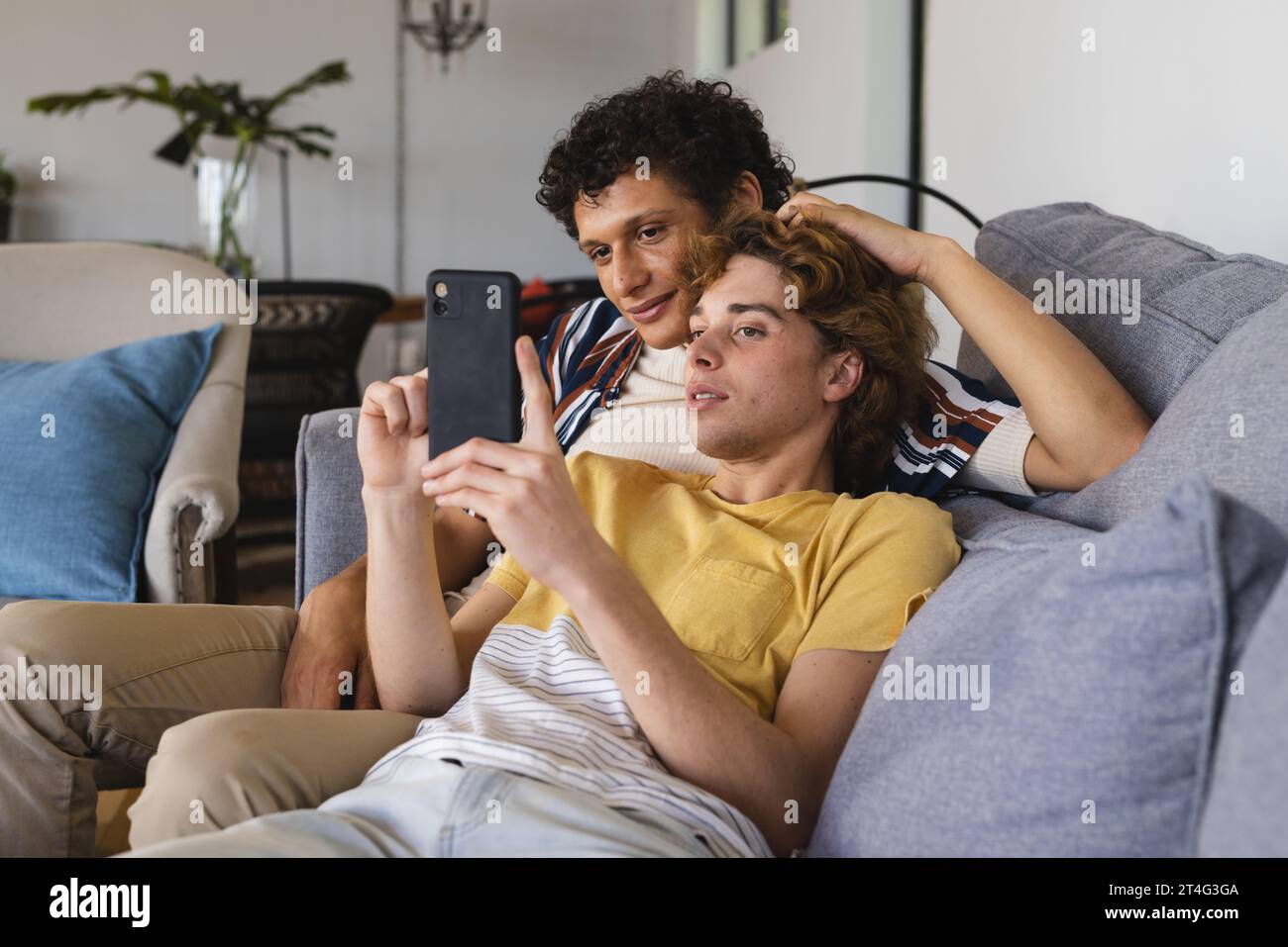 Heureux couple masculin gay diversifié utilisant smartphone et embrassant à la maison Banque D'Images