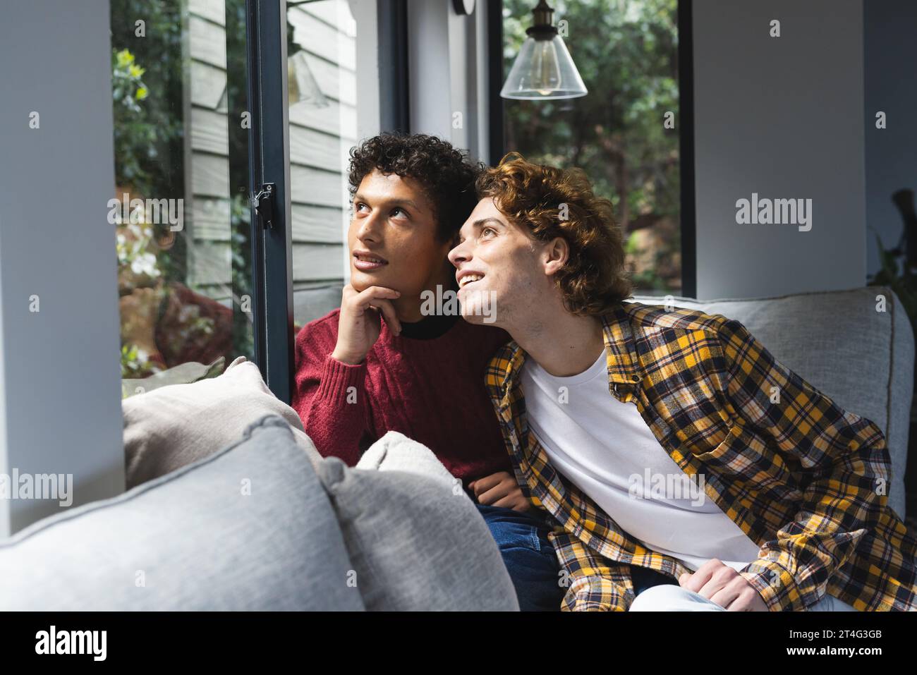 Heureux couple masculin gay diversifié embrassant et regardant par la fenêtre à la maison Banque D'Images