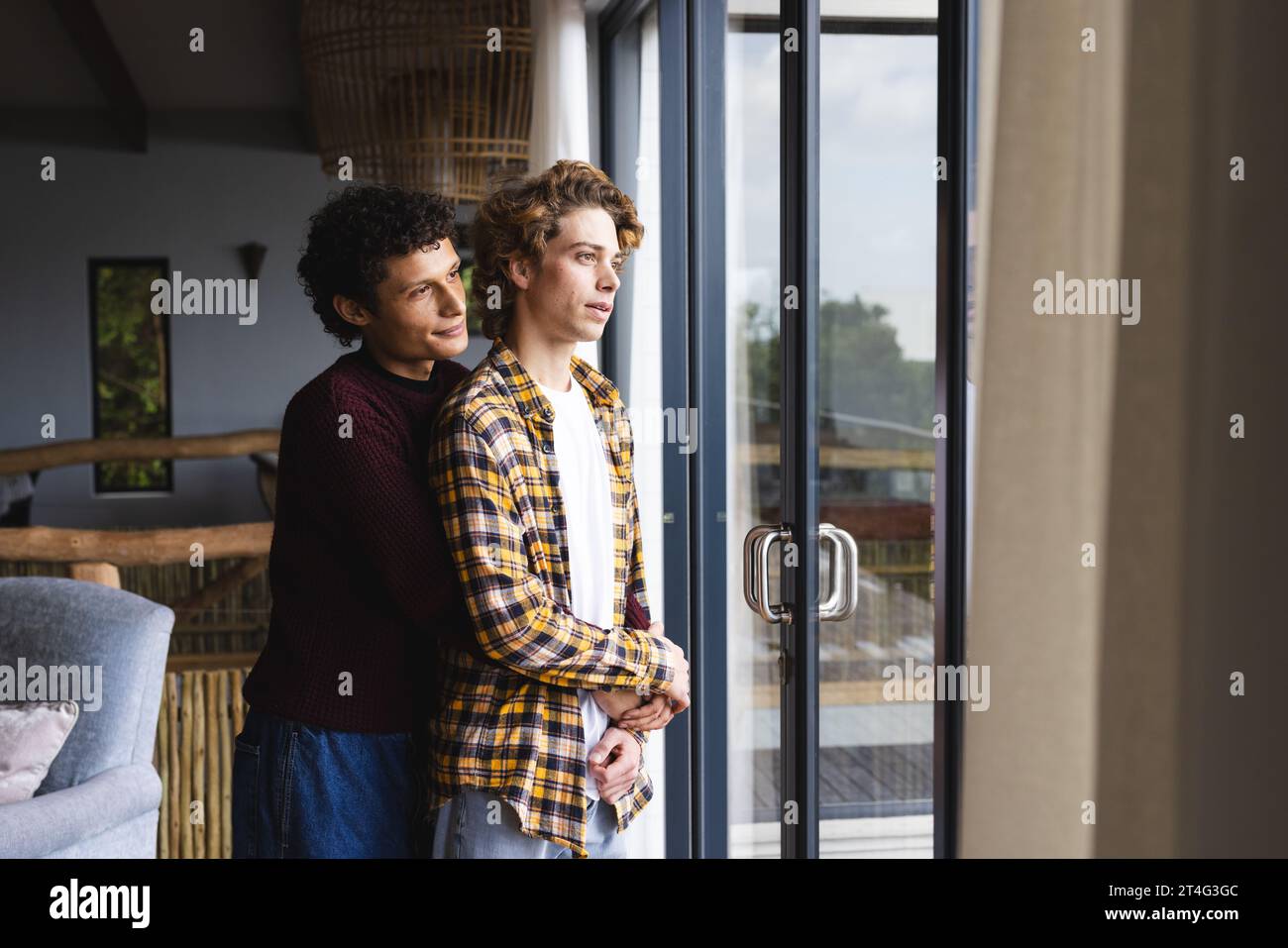 Heureux couple masculin gay diversifié embrassant et regardant par la fenêtre à la maison Banque D'Images
