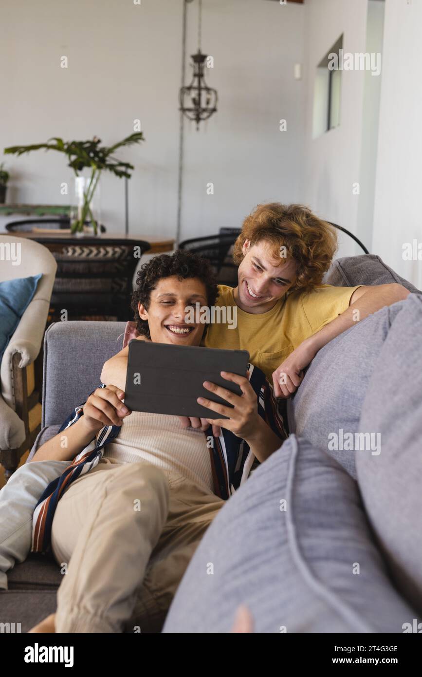 Heureux couple masculin gay diversifié utilisant la tablette et embrassant à la maison Banque D'Images