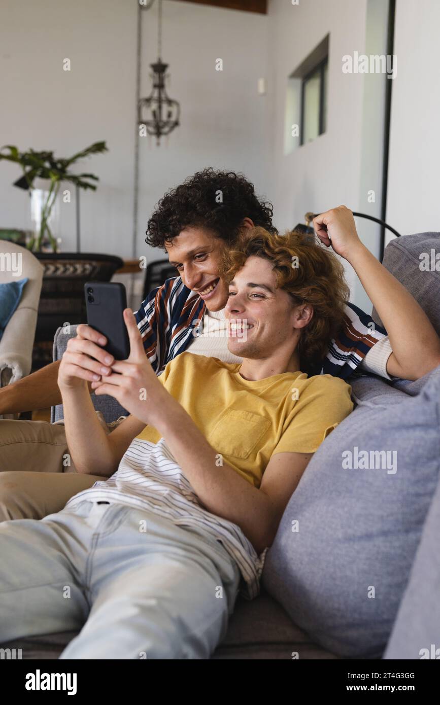 Heureux couple masculin gay diversifié utilisant smartphone et embrassant à la maison Banque D'Images