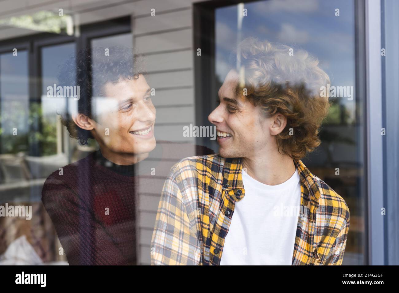 Heureux et diversifié couple masculin gay embrassant et se regardant à la fenêtre à la maison Banque D'Images