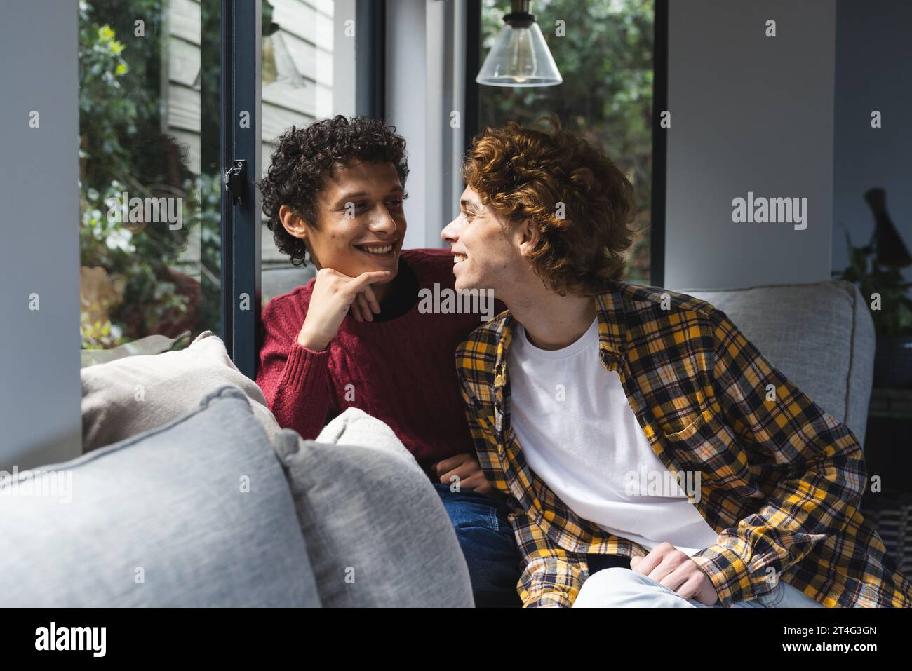 Heureux et diversifié couple masculin gay embrassant et se regardant à la fenêtre à la maison Banque D'Images