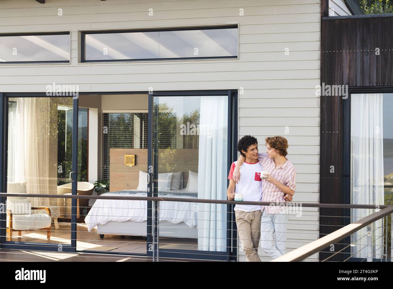 Heureux et diversifié couple masculin gay embrassant et tenant des tasses de café sur le balcon Banque D'Images