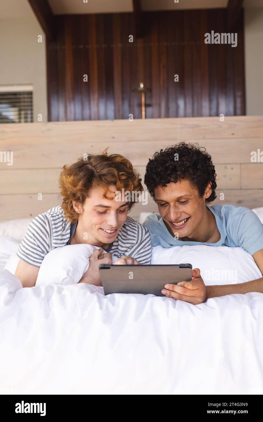 Heureux couple masculin gay diversifié couché sur le lit et utilisant tablette à la maison Banque D'Images