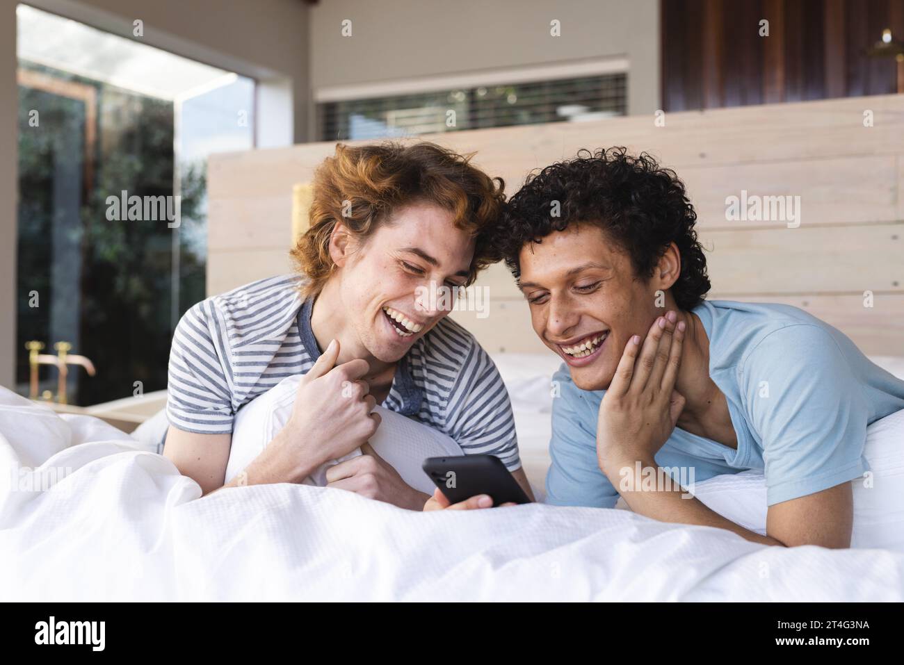 Heureux couple masculin gay diversifié couché sur le lit et utilisant smartphone à la maison Banque D'Images