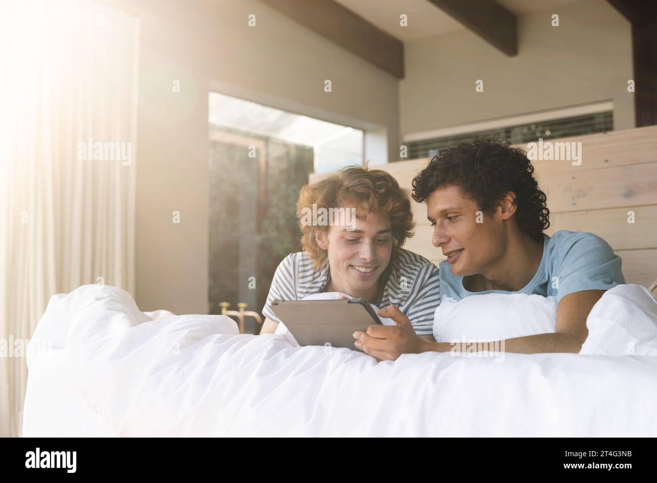 Heureux couple masculin gay diversifié couché sur le lit et utilisant tablette à la maison Banque D'Images