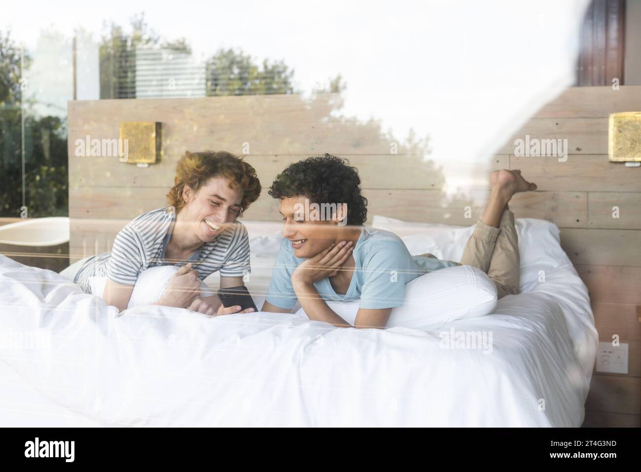 Heureux couple masculin gay diversifié couché sur le lit et utilisant smartphone à la maison Banque D'Images