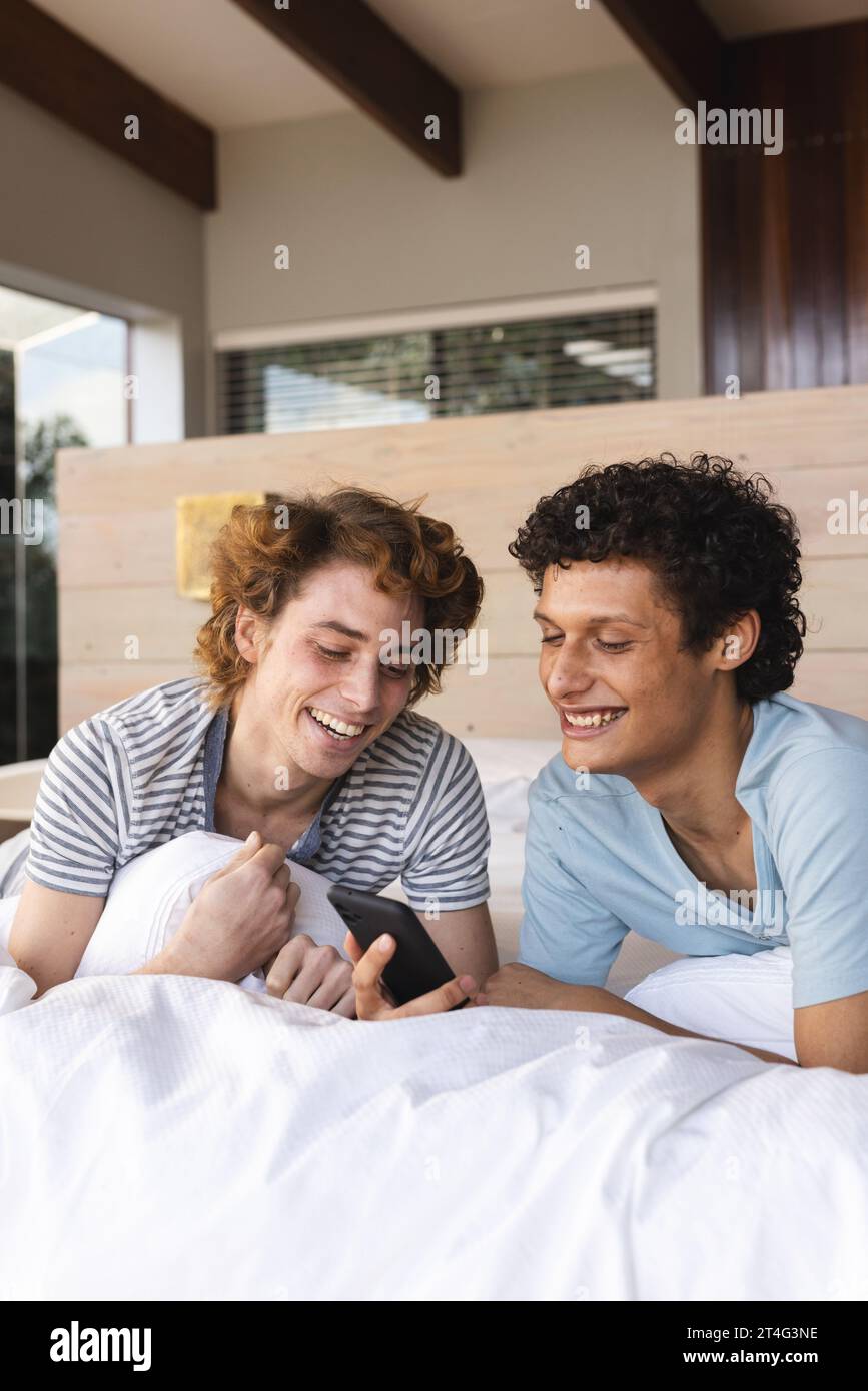 Heureux couple masculin gay diversifié couché sur le lit et utilisant smartphone à la maison Banque D'Images