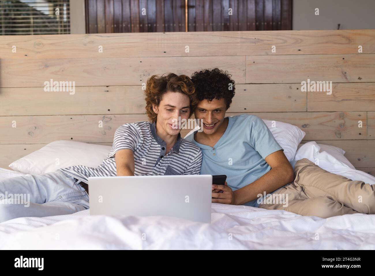 Heureux couple masculin gay diversifié couché sur le lit et utilisant un ordinateur portable à la maison Banque D'Images