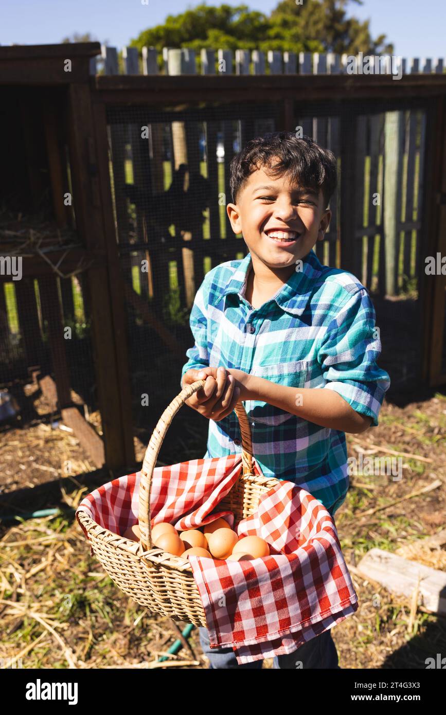 Portrait de garçon biracial heureux tenant le panier avec des œufs devant le poulailler Banque D'Images