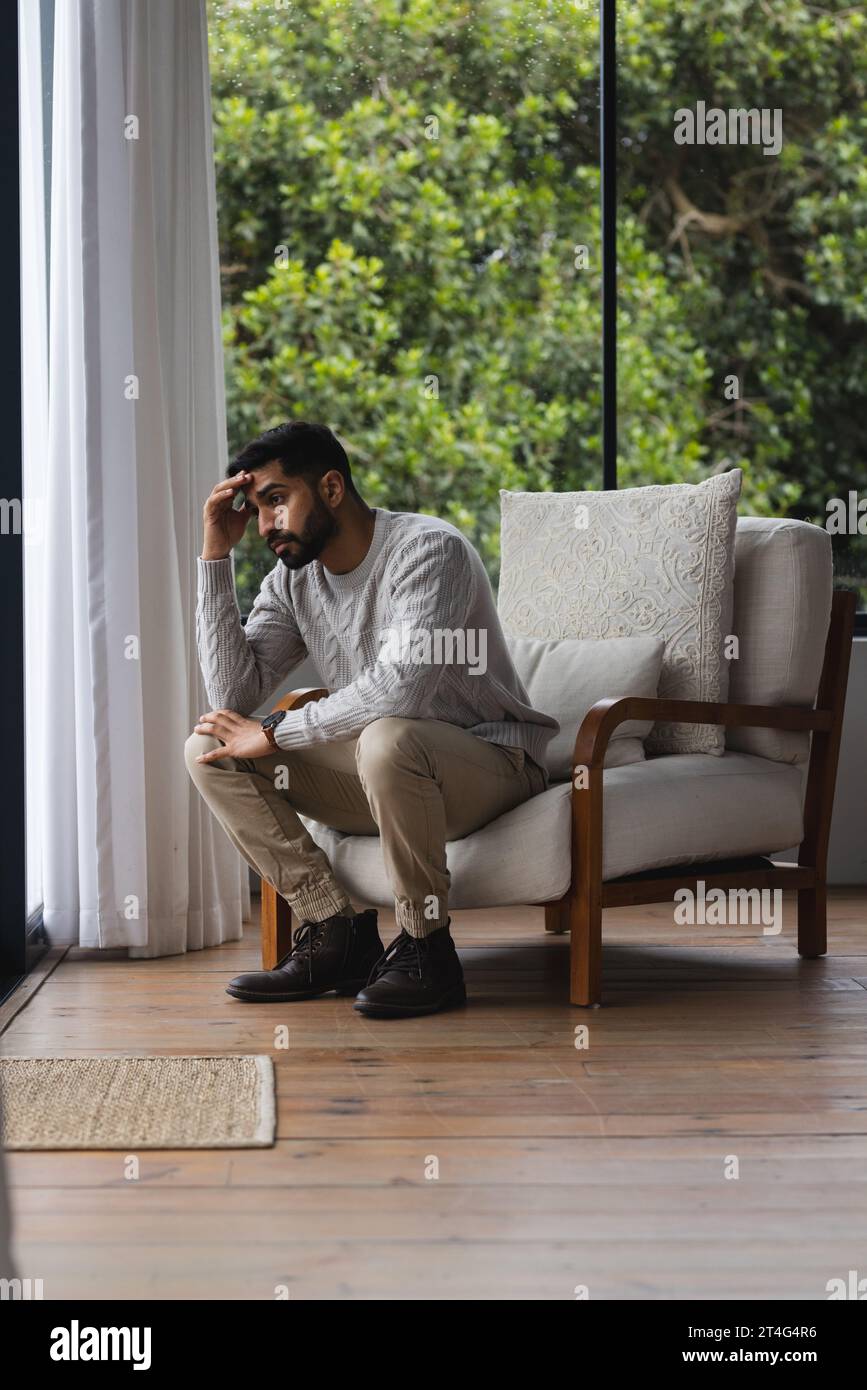 Homme biracial inquiet assis sur le fauteuil et regardant par la fenêtre dans le salon ensoleillé Banque D'Images