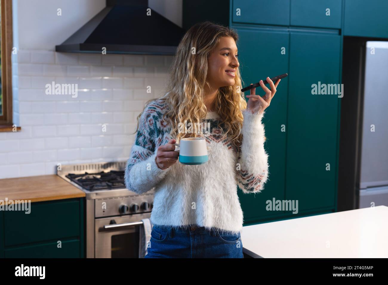 Heureuse femme caucasienne tenant le café et parlant sur smartphone debout dans la cuisine Banque D'Images