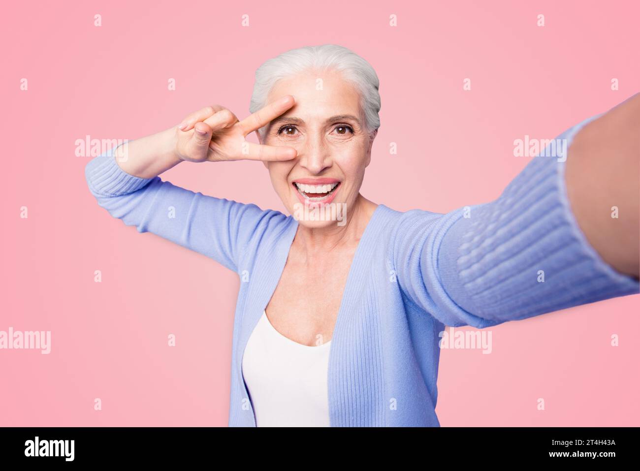 Une dame âgée fait un selfie sur l'appareil photo d'un smartphone faisant un v-sign près des yeux isolés sur fond violet clair avec un espace de copie pour le texte Banque D'Images