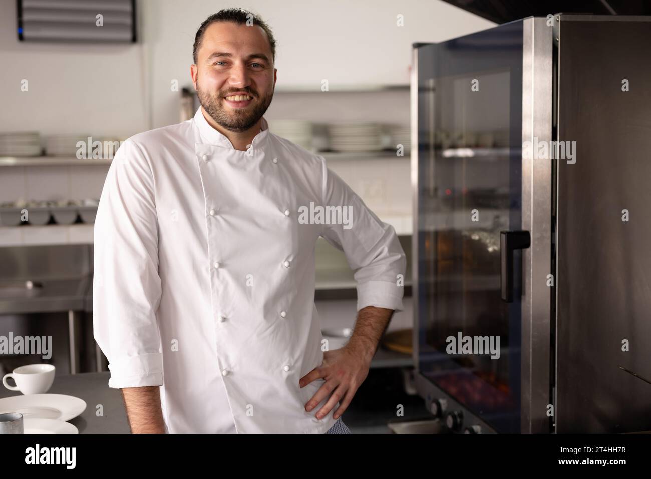 Portrait d'un chef caucasien souriant avec la main sur la hanche debout dans la cuisine à l'école de cuisine Banque D'Images