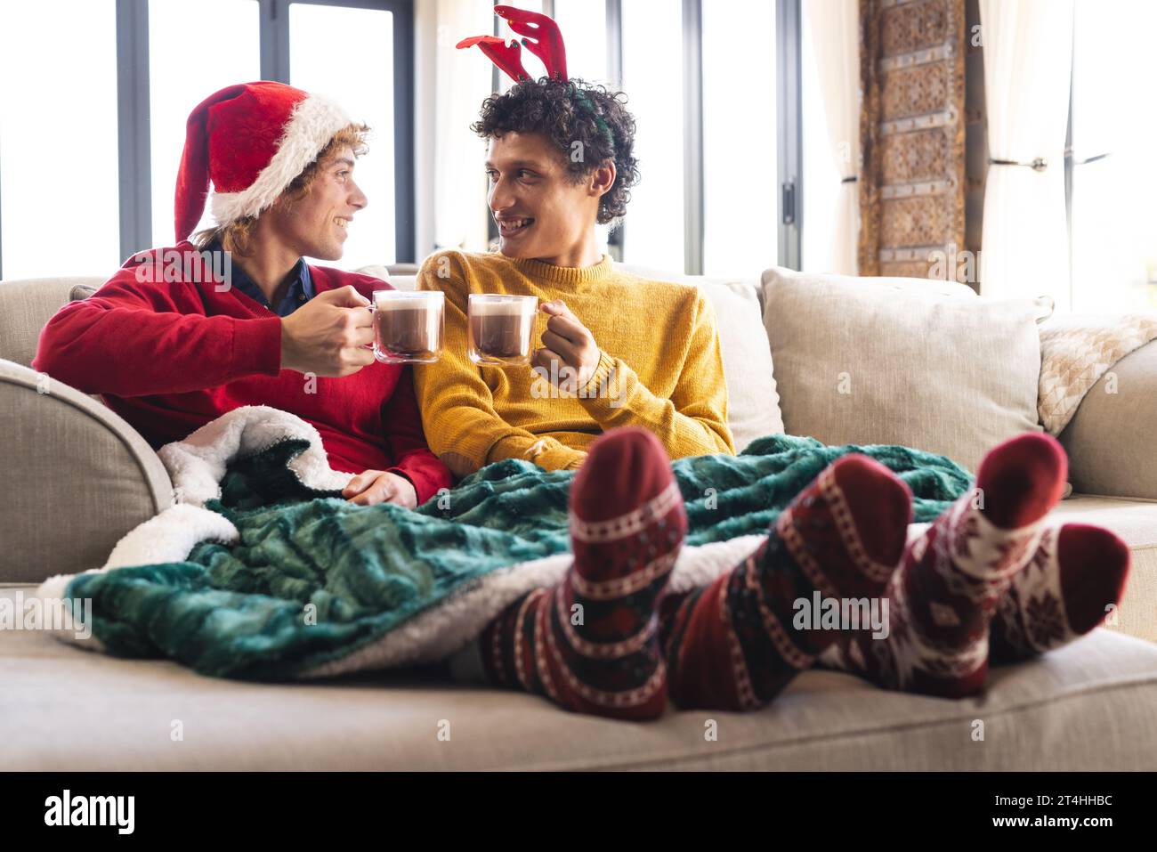 Heureux couple masculin gay diversifié buvant du cacao et portant des vêtements de noël à la maison Banque D'Images