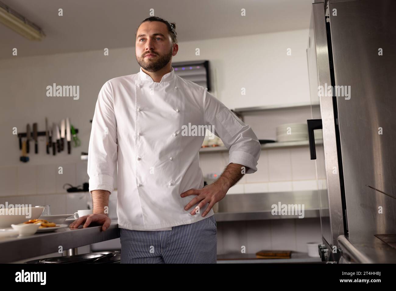 Chef masculin caucasien réfléchi avec la main sur la hanche debout dans la cuisine à l'école de cuisine Banque D'Images
