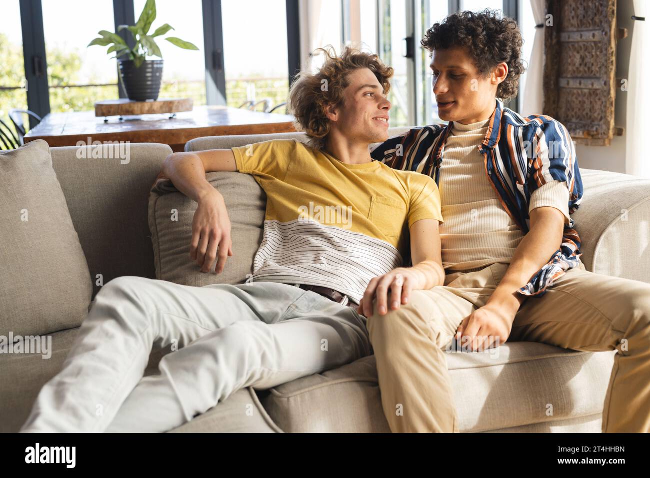 Heureux couple masculin gay diversifié assis sur le canapé et embrassant à la maison Banque D'Images