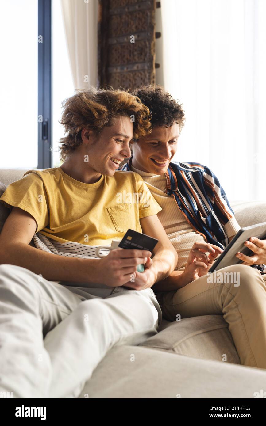 Heureux couple masculin gay diversifié utilisant tablette avec carte de crédit à la maison Banque D'Images