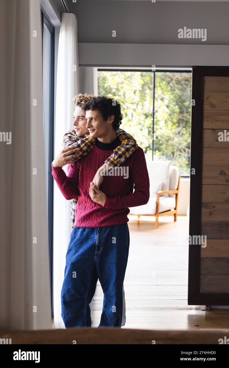 Heureux couple masculin gay diversifié embrassant et regardant par la fenêtre à la maison Banque D'Images