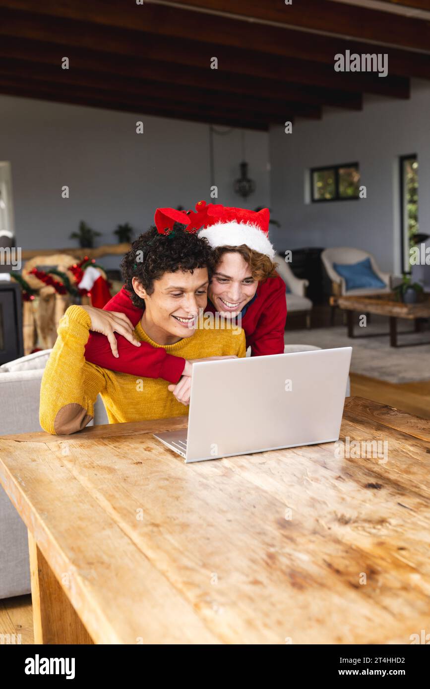 Heureux couple masculin gay diversifié embrassant porter des chapeaux de noël et ayant un appel vidéo à la maison Banque D'Images