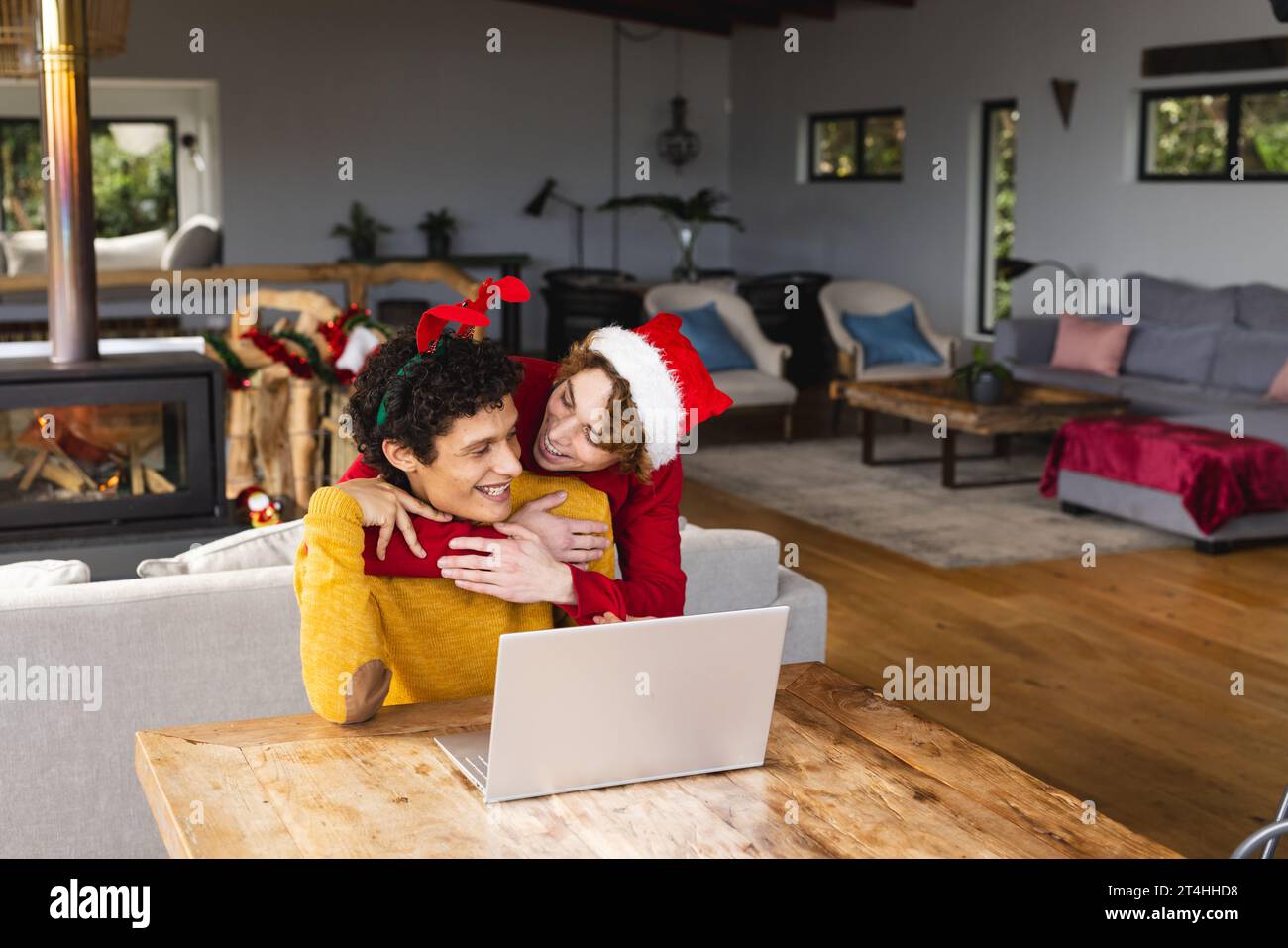 Heureux couple masculin gay diversifié embrassant porter des chapeaux de noël et ayant un appel vidéo à la maison Banque D'Images
