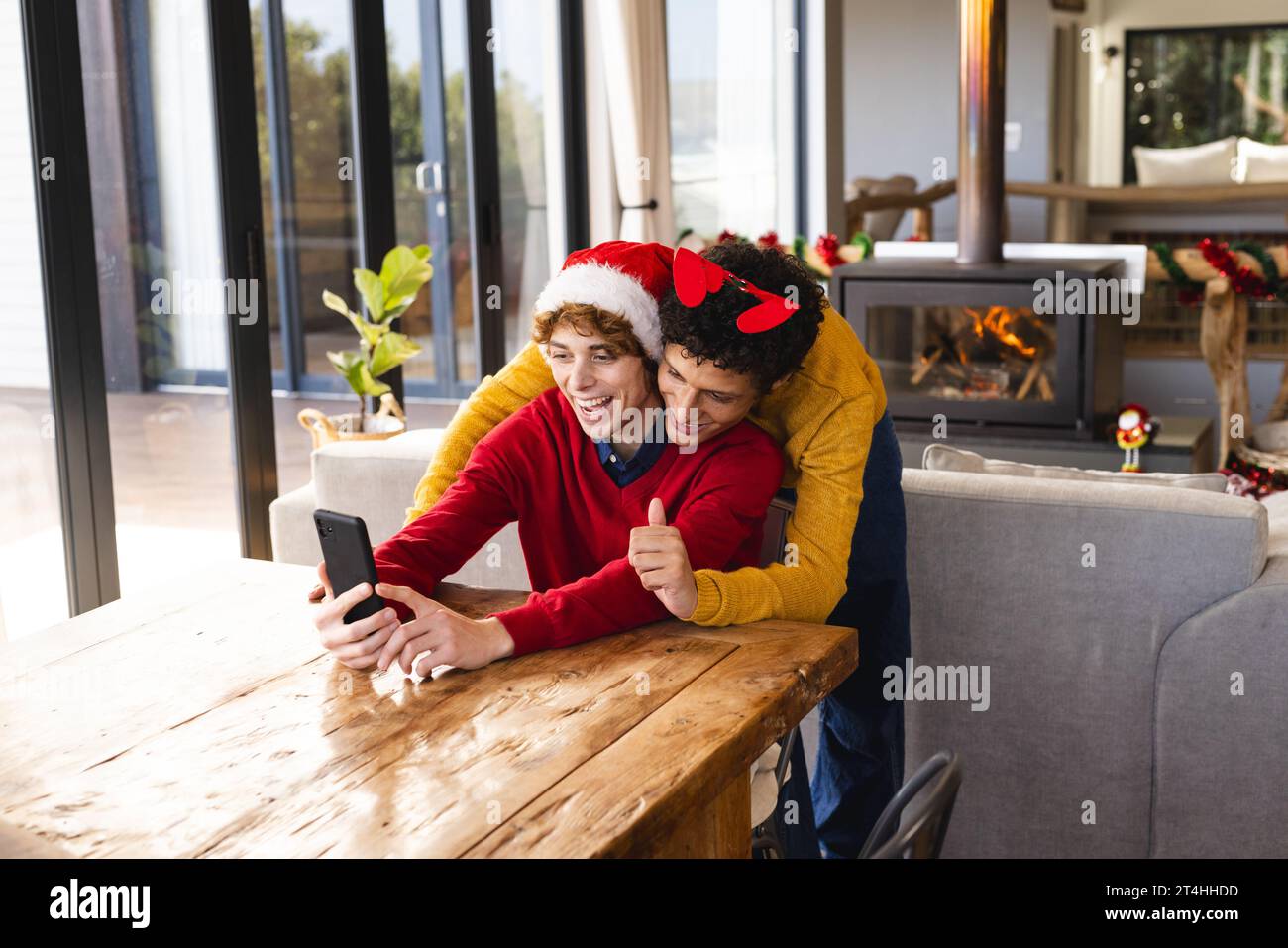 Heureux couple masculin gay diversifié embrassant porter des chapeaux de noël et ayant un appel vidéo à la maison Banque D'Images