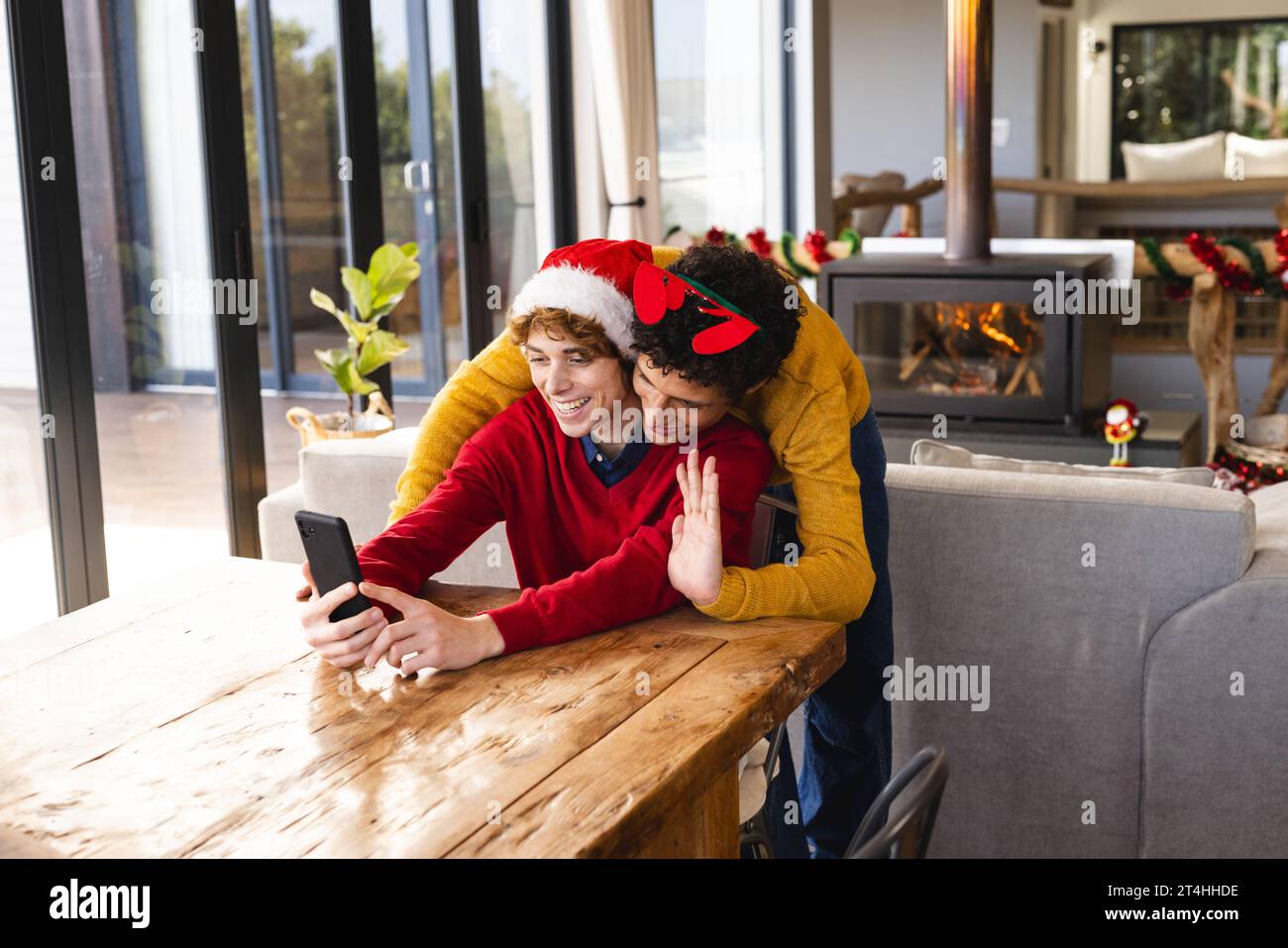 Heureux couple masculin gay diversifié embrassant porter des chapeaux de noël et ayant un appel vidéo à la maison Banque D'Images