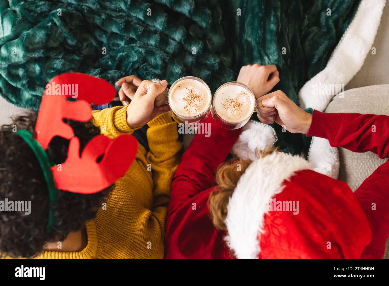 Couple masculin gay diversifié tenant des verres de cacao et portant des chapeaux de noël à la maison Banque D'Images